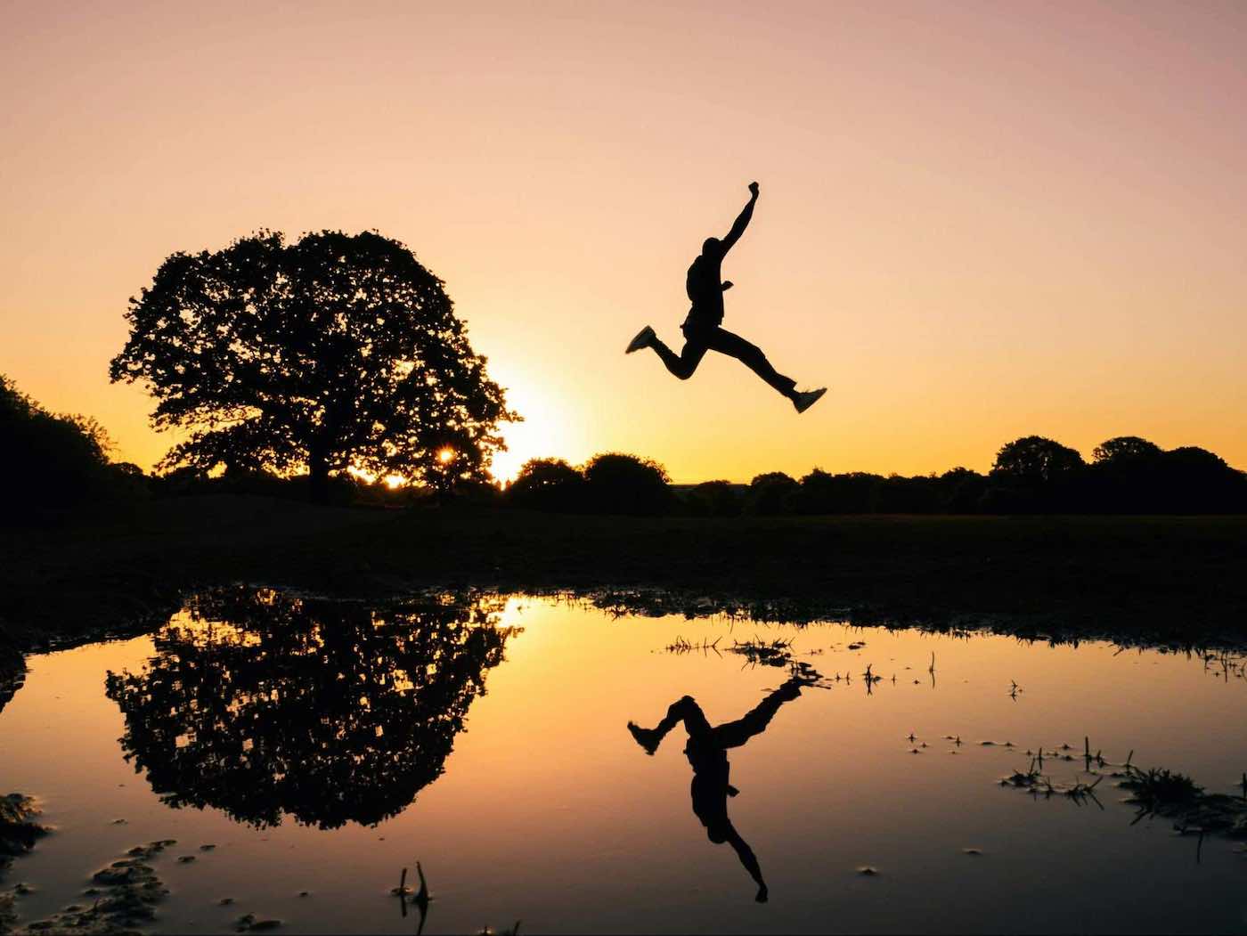 Silhouette of a person leaping across a reflective pond at sunset, with a large tree and vibrant orange sky in the background.