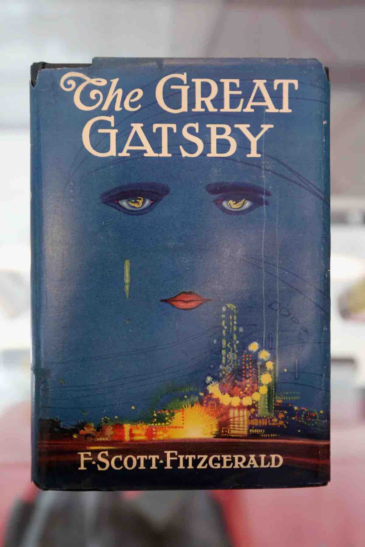 Vintage book cover of ’The Great Gatsby’ featuring a large blue face with piercing eyes and a vibrant cityscape along the bottom.