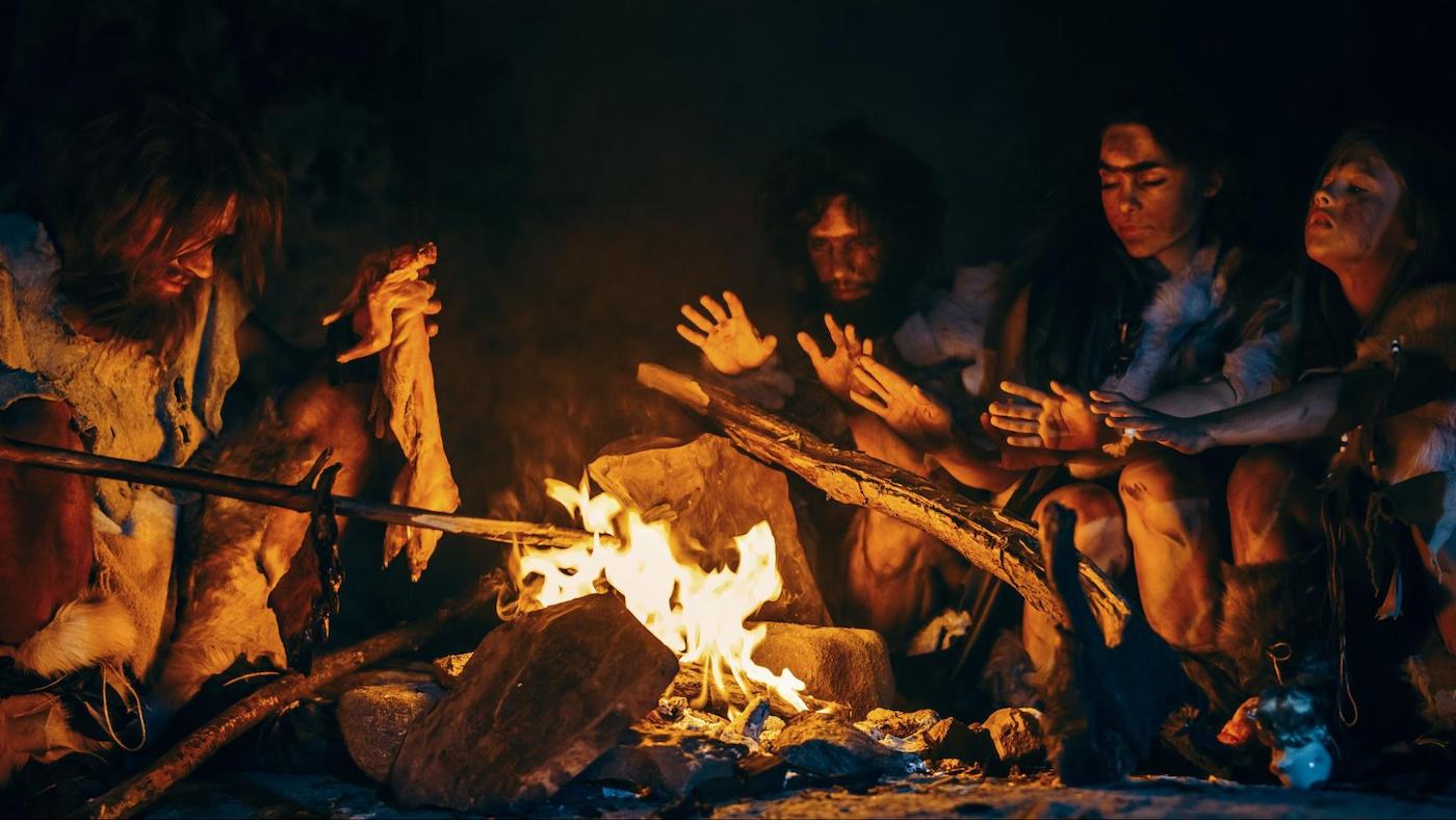 Prehistoric humans sitting around a campfire, cooking meat and warming their hands in a cave setting.