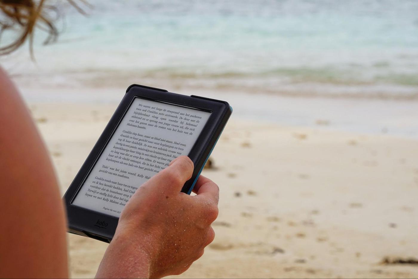 Person reading an e-book on a tablet while relaxing on a sandy beach by the ocean.