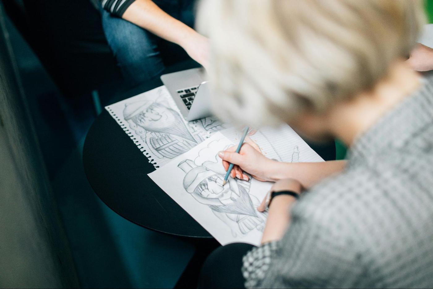 Artist sketching detailed character drawings on paper during a collaborative creative session, with sketches and a laptop on the table.