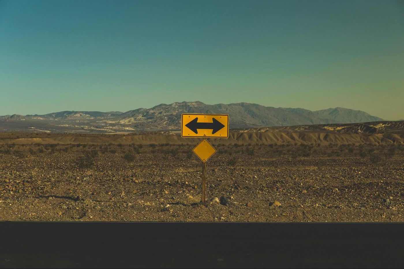 Yellow directional road sign with arrows pointing left and right in a desolate desert landscape with distant mountains.