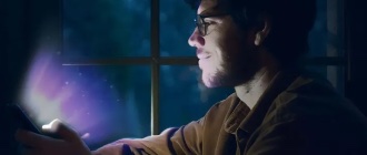 Man looking at a glowing phone screen with a smile, illuminated by soft purple light in a dimly lit room.