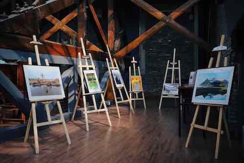 Art gallery exhibition featuring landscape paintings displayed on wooden easels in a rustic setting.