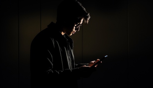 Young person standing in a dark room, intently reading something on his smartphone.